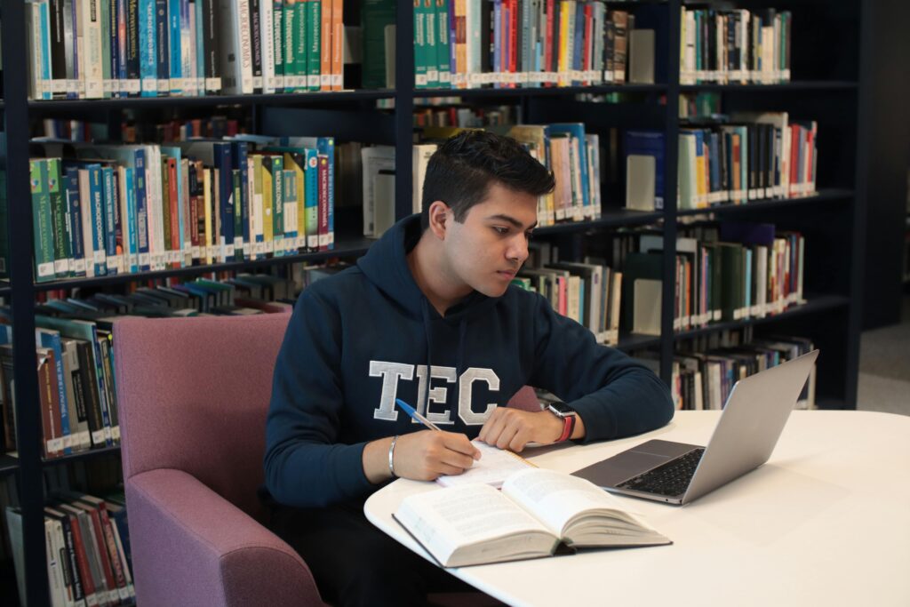 A man in a hoodie studies in a library, using books and a laptop on a table.
