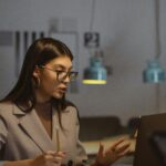 A young businesswoman working late at her desk, engaged in an online meeting with a laptop, notebook, and smartphone.