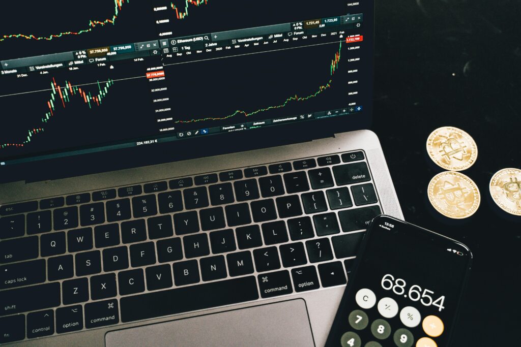 A laptop with trading charts, smartphone calculator, and bitcoin coins depicting cryptocurrency trading.