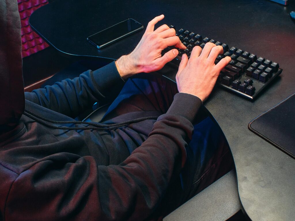 Person in hoodie typing on a keyboard in a dark room, focusing on hacking activities.